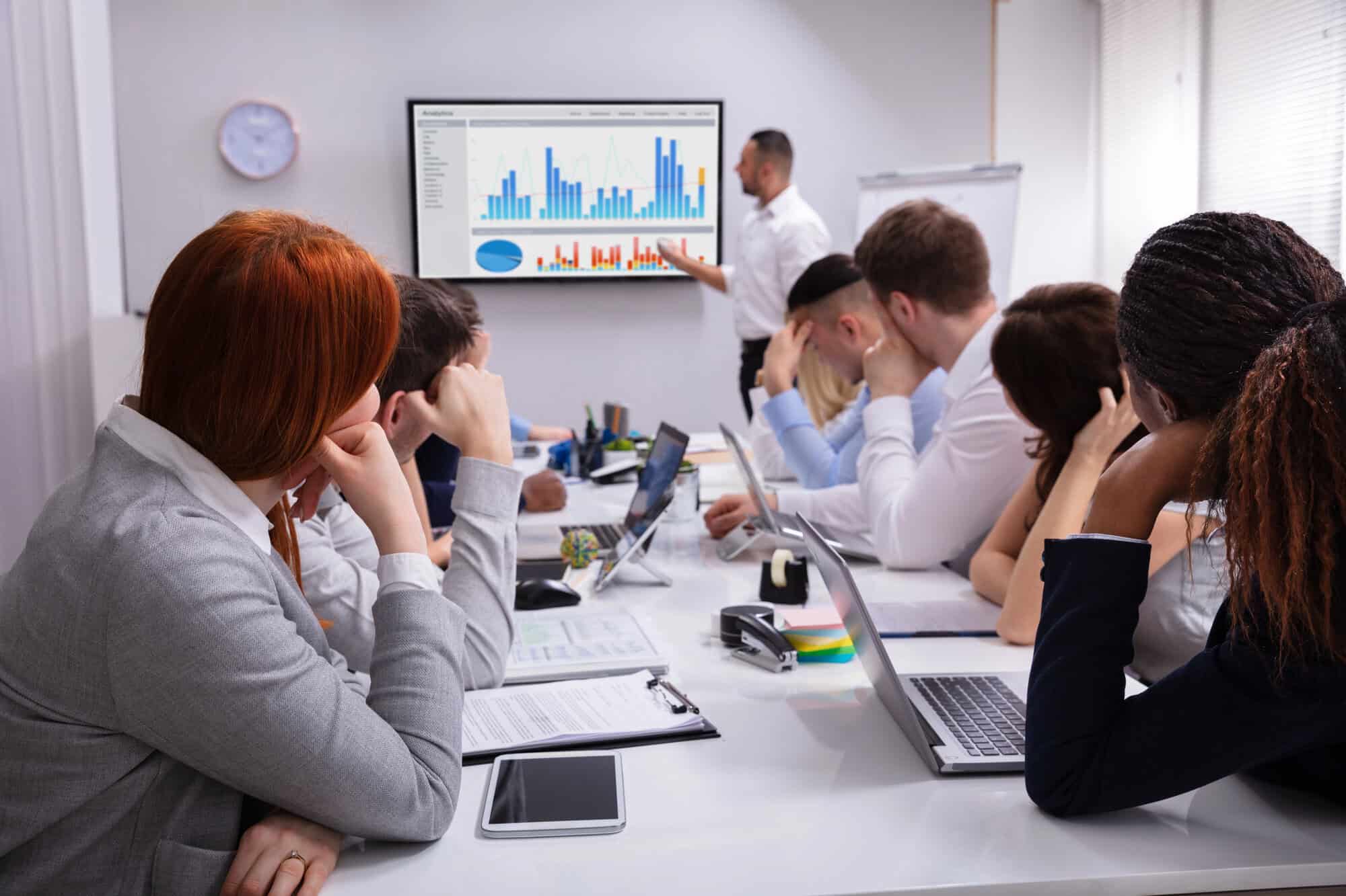 A business meeting in progress with a man presenting a bar graph on a screen. Five colleagues sit around a table, some with laptops and notes.