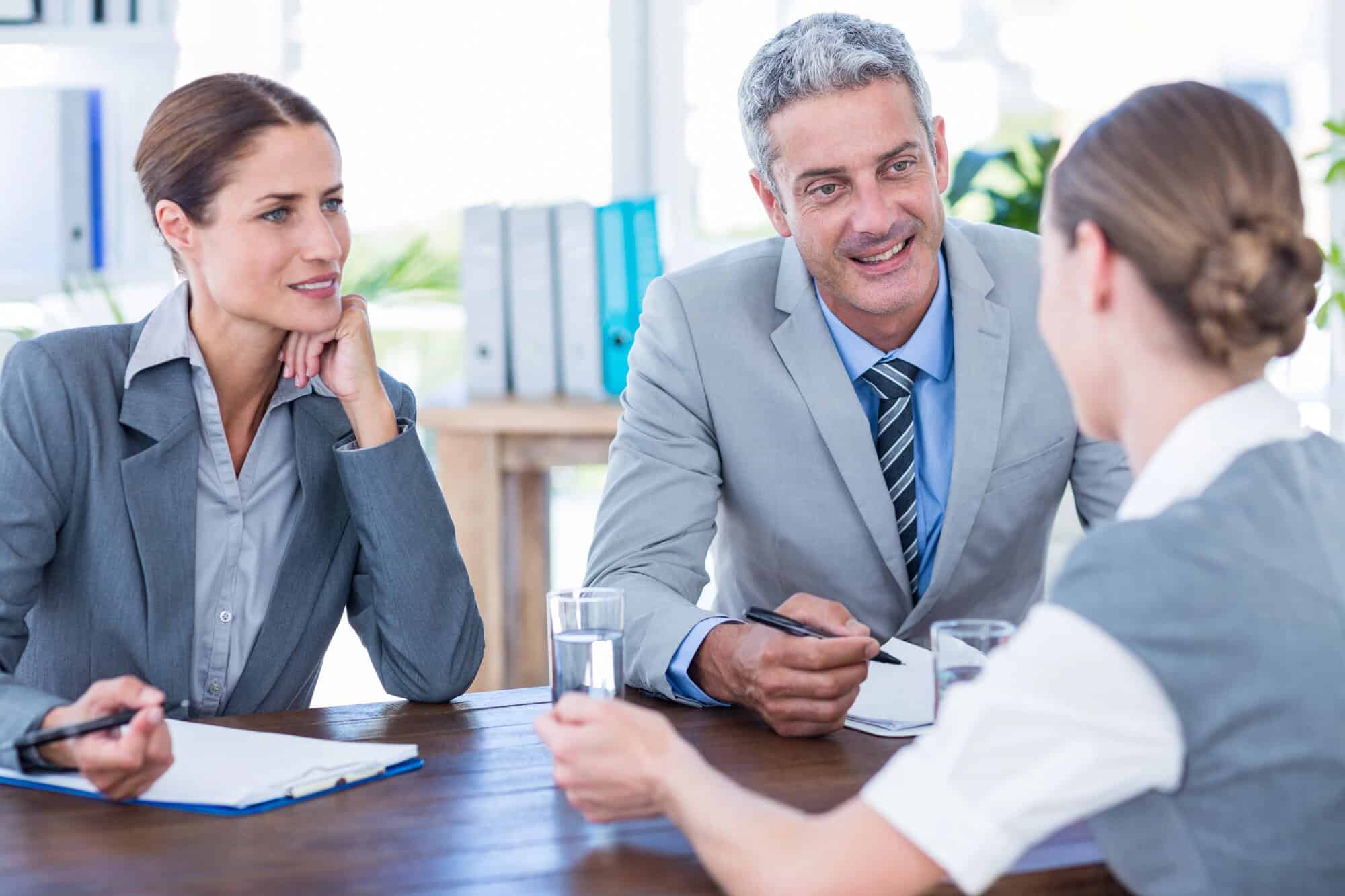 Three professionals in business attire engage in a conversation around a table in a bright office setting, with notepads and glasses of water visible on the table.