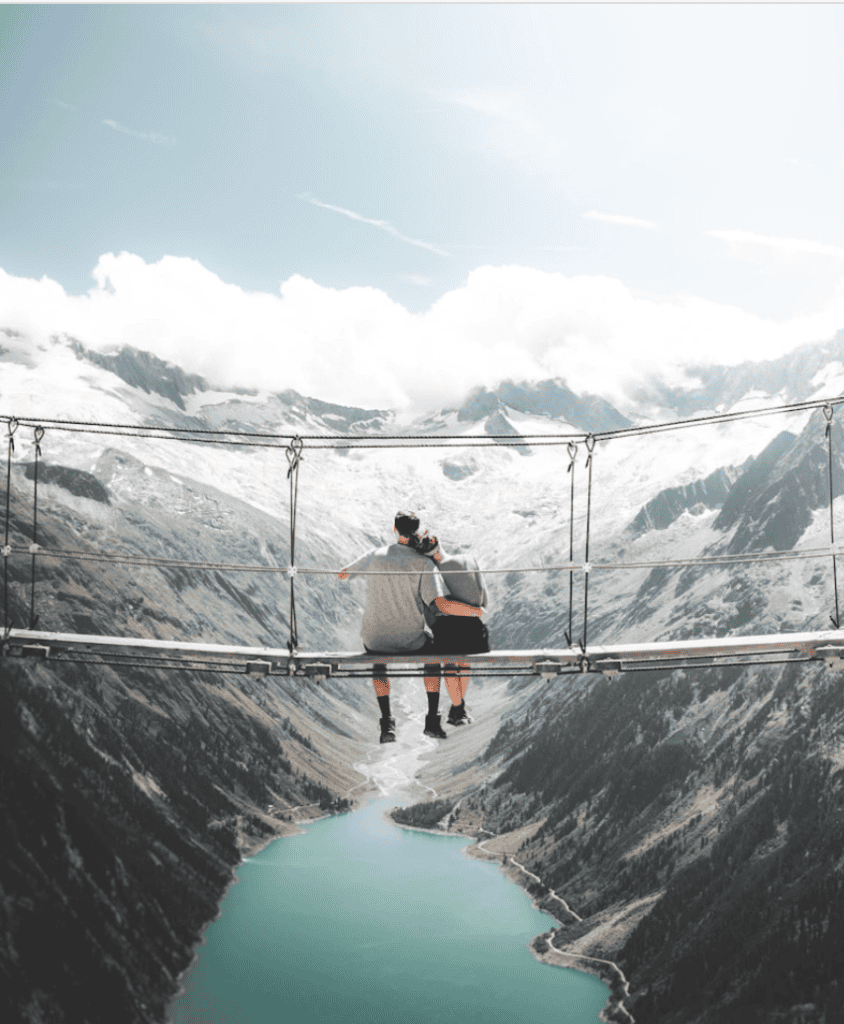 Two people sit on a suspension bridge, overlooking a scenic view of mountains and a river below.