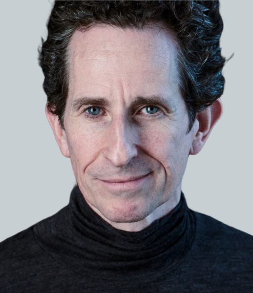 Man with short, curly dark hair wearing a dark turtleneck, looking directly at the camera against a plain background.