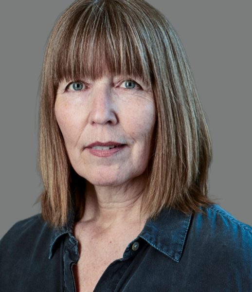 A person with straight, shoulder-length hair and bangs, wearing a dark shirt, against a plain background.