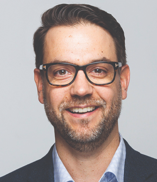 A person with short dark hair, glasses, and a beard is smiling. They are wearing a dark jacket and a light-colored shirt, set against a plain background.