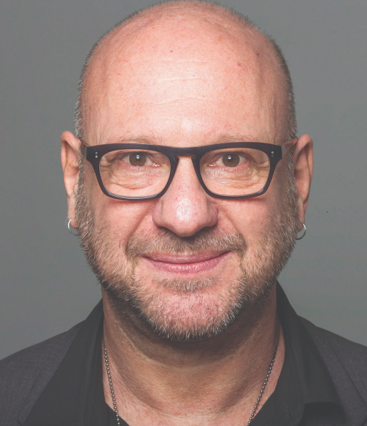A man with glasses, earrings, and a beard, wearing a dark shirt, smiles at the camera against a plain background.