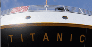 Close-up view of the Titanic's bow, showing the ship's name and a red warning sign about the vessel's triple screws, with part of the deck and railing visible.
