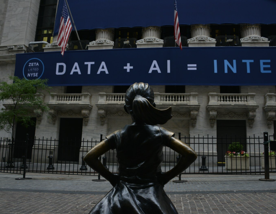 A bronze statue of a girl stands facing a building displaying a large banner that reads "DATA + AI =" with American flags above.