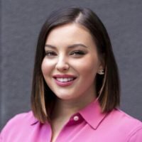 A woman with shoulder-length dark hair and wearing a pink shirt smiles at the camera against a grey background.